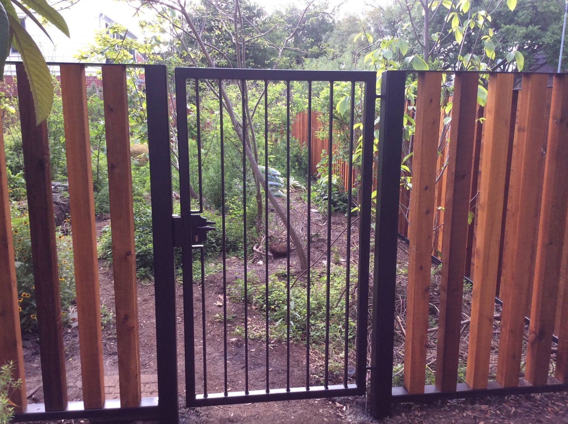 Black metal gate in front of a lush garden path, with wooden fence panels on both sides.