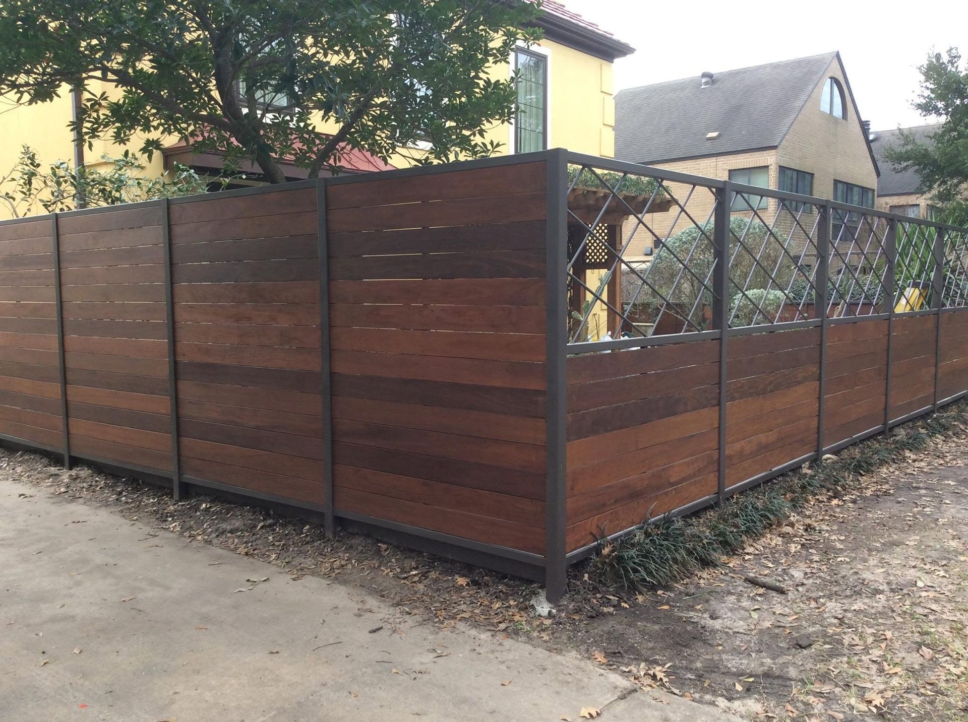 Wooden fence enclosing a corner yard beside houses, with metal gate and dark wood panels
