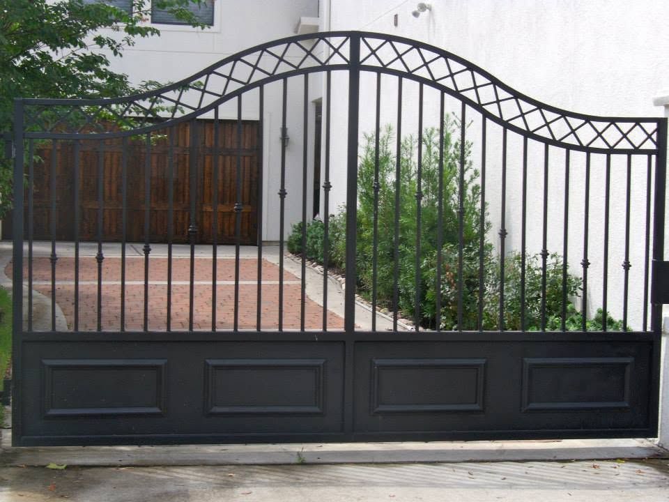 Black metal driveway gate with arched top in front of a house and garden