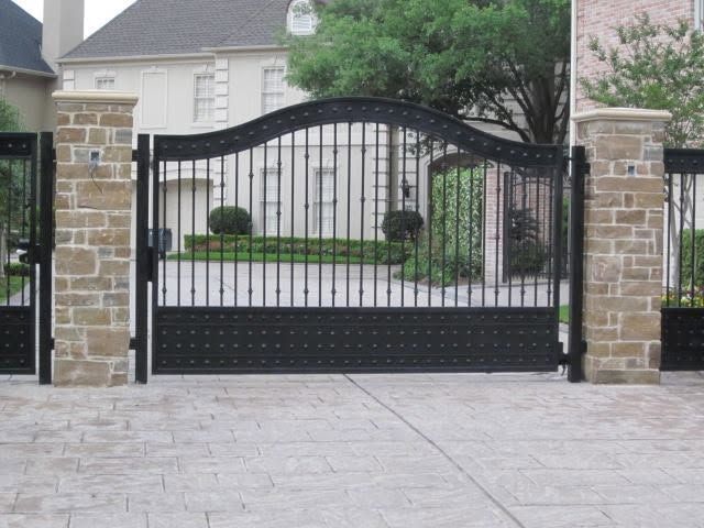 Black wrought-iron driveway gate between stone pillars in front of a house