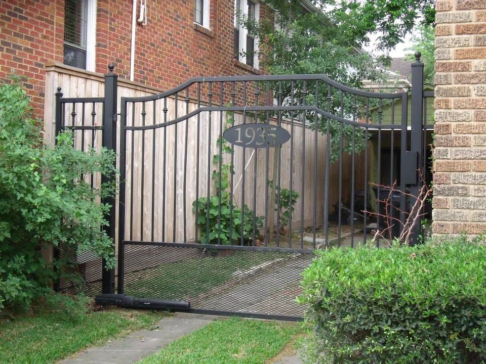 Black wrought-iron gate between brick walls with house number 1976 and greenery beyond.