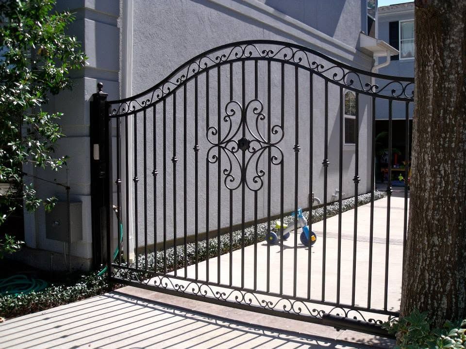 Decorative black wrought-iron gate across a driveway beside a house and tree