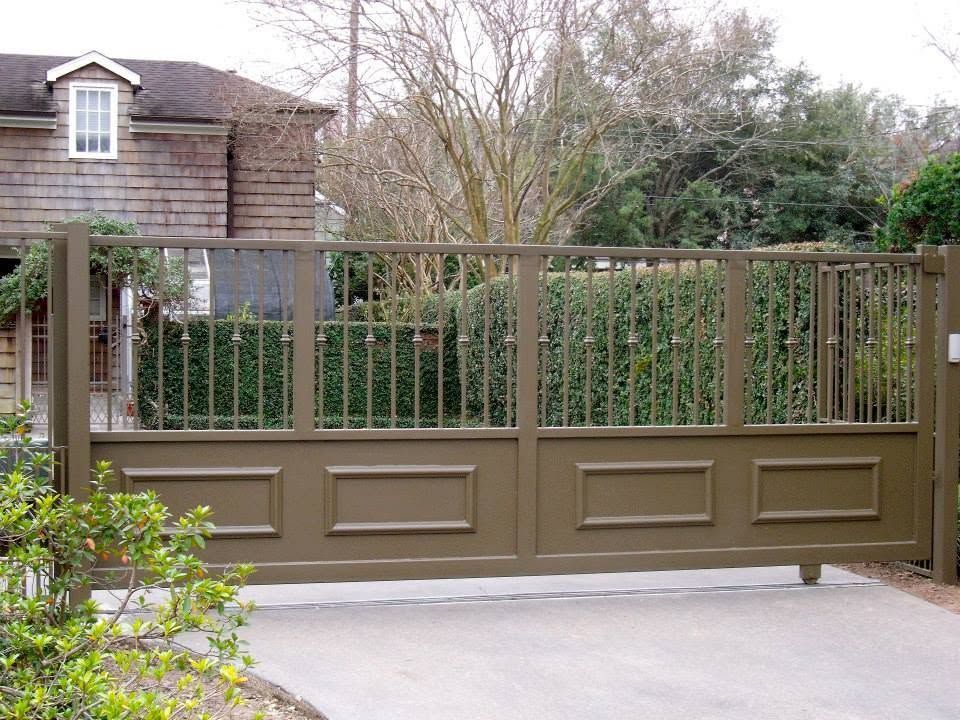 Closed brown driveway gate with vertical bars in front of a house and hedge