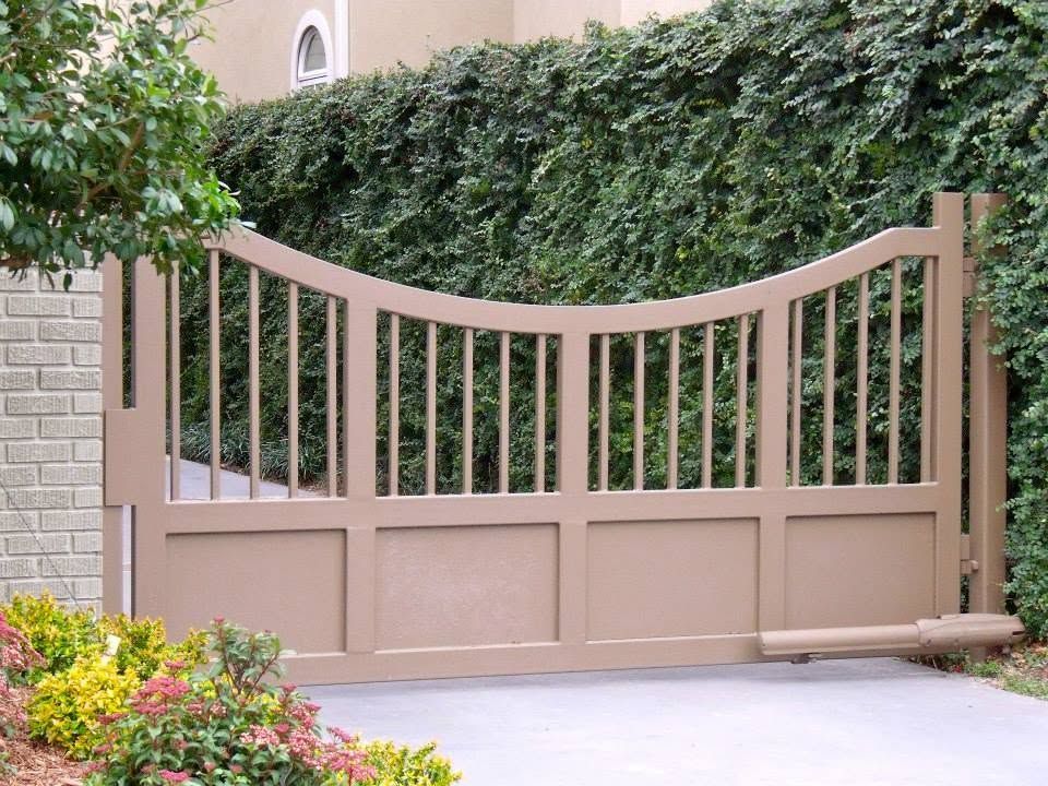 Tan curved beige driveway gate with vertical slats in front of dense green hedges