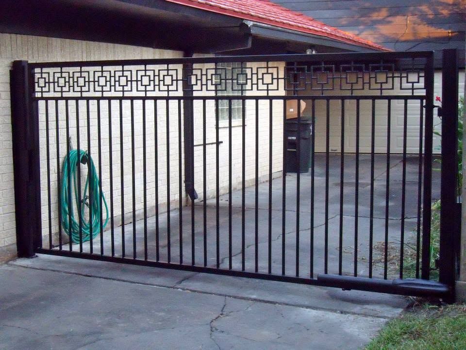 Black metal driveway gate with geometric panels in a concrete courtyard, attached to a house with a red roof.