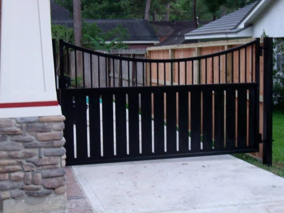 Black metal driveway gate with vertical slats beside a stone-sided house and concrete driveway