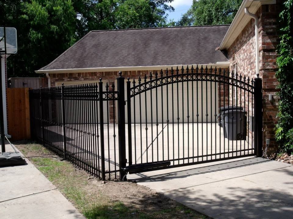 Black metal driveway gate in front of a tan garage and brick house, beside a sidewalk.