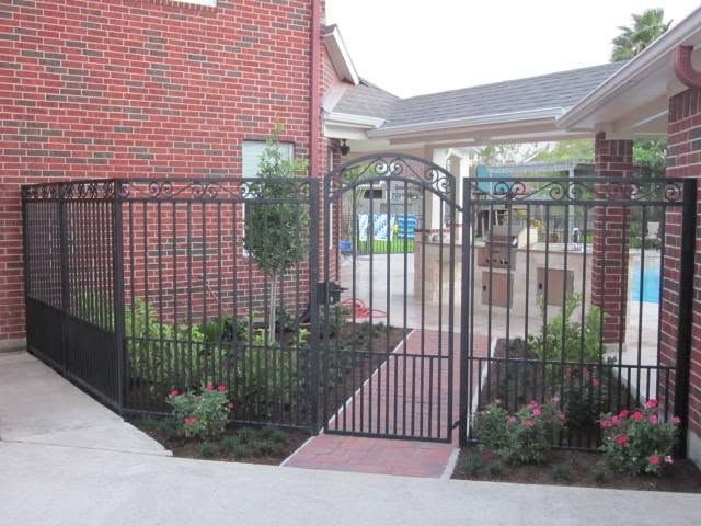 Black metal gate entrance between brick buildings with flower beds and a walkway