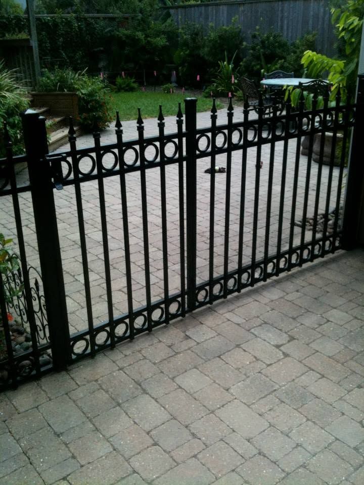 Black metal garden gate across a brick patio with a backyard and patio furniture behind it