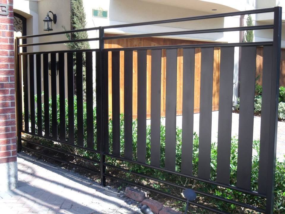 Black metal gate along a brick walkway beside a building entrance and wooden fence panels.