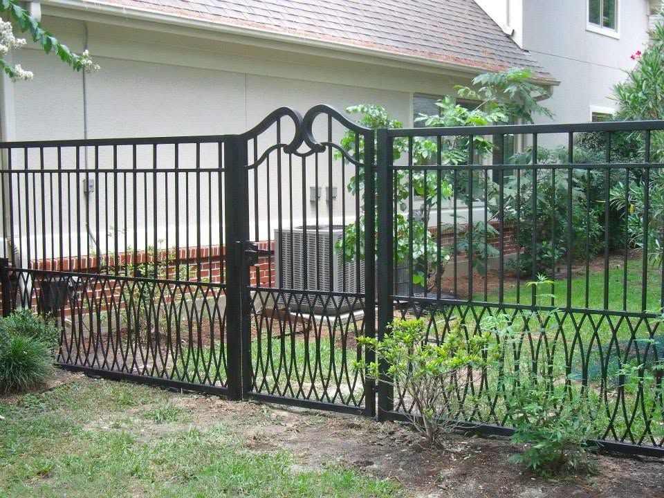 Black metal garden gate and fence beside a house, with shrubs and a grassy yard.