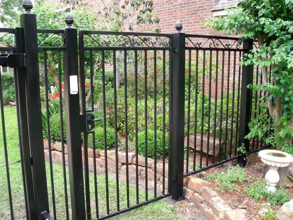 Black metal garden gate beside a brick house, opening to a leafy path.