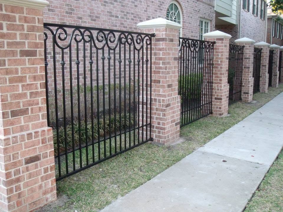Black wrought-iron fence between brick columns beside a sidewalk and apartment building