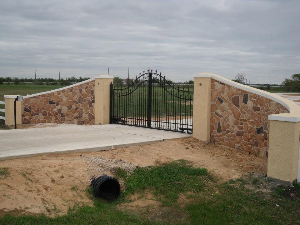 Stone entrance with black wrought-iron gate and curved walls over a small drainage culvert.