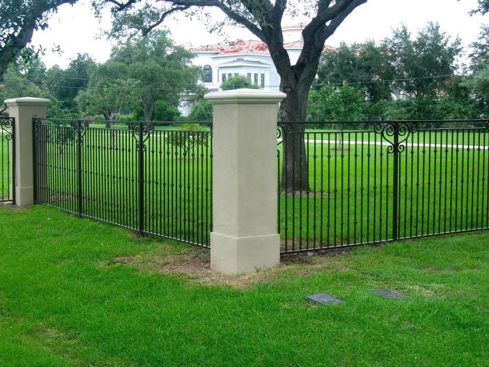 Black iron fence between stone pillars around a green lawn with a large tree and house in the background