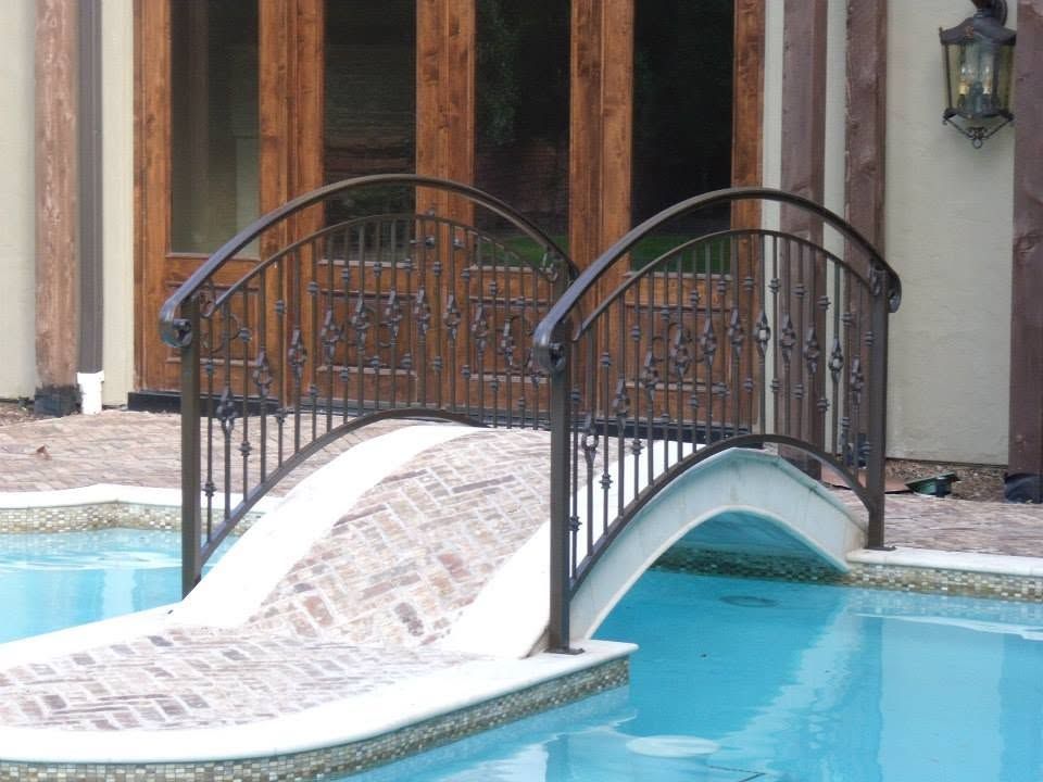 Small arched footbridge with black railings over a turquoise pool beside a building