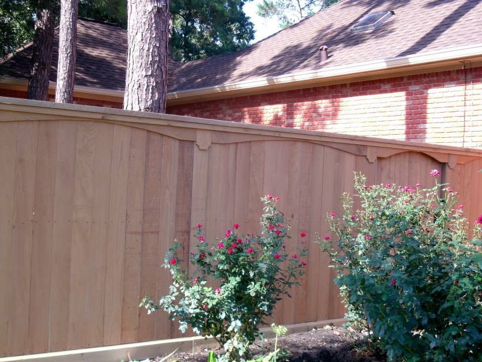 Wooden privacy fence beside a brick house, with green shrubs and tall pine trees in the background