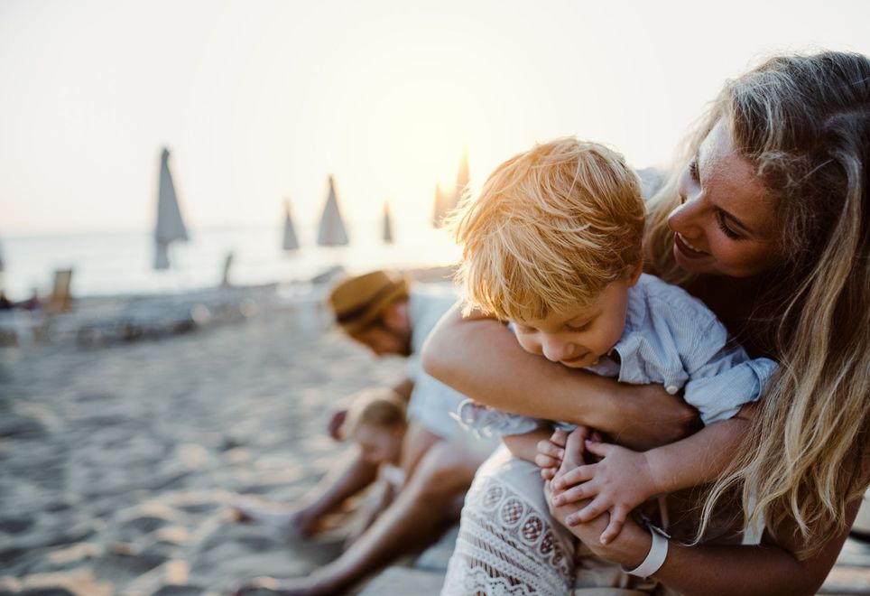 Mother hugging a child on a beach at sunset, with a person and another child in the blurred background.