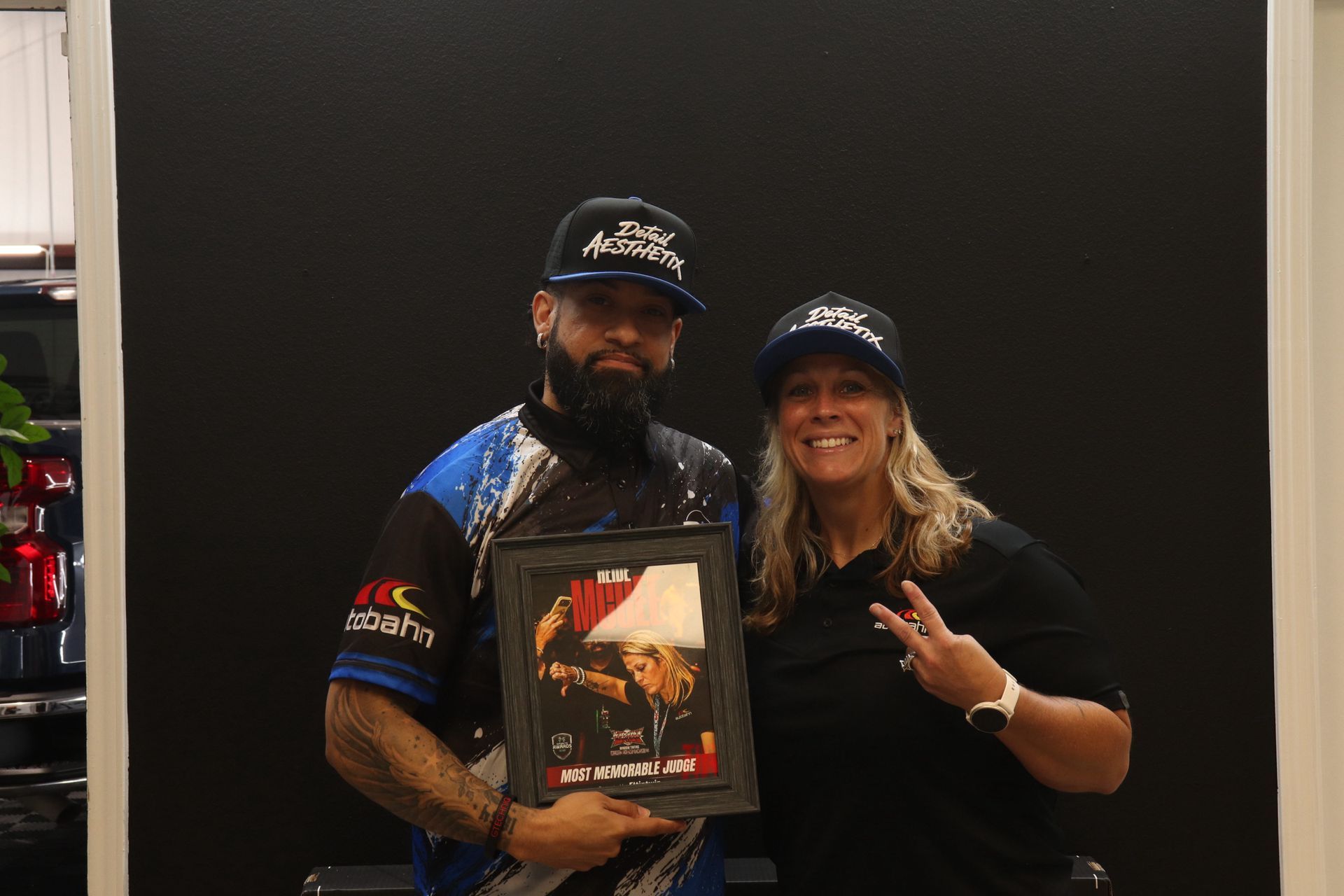 Two people posing indoors, one holding a framed award, both wearing black caps and smiling