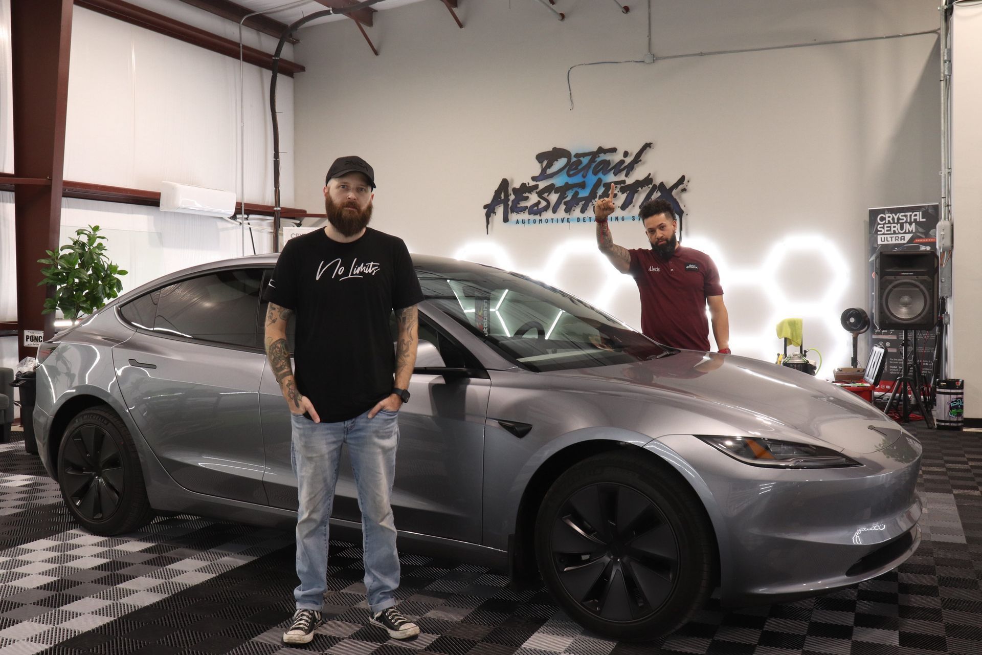 Two men stand beside a gray sports sedan in a garage, with automotive lights and signage in the background.