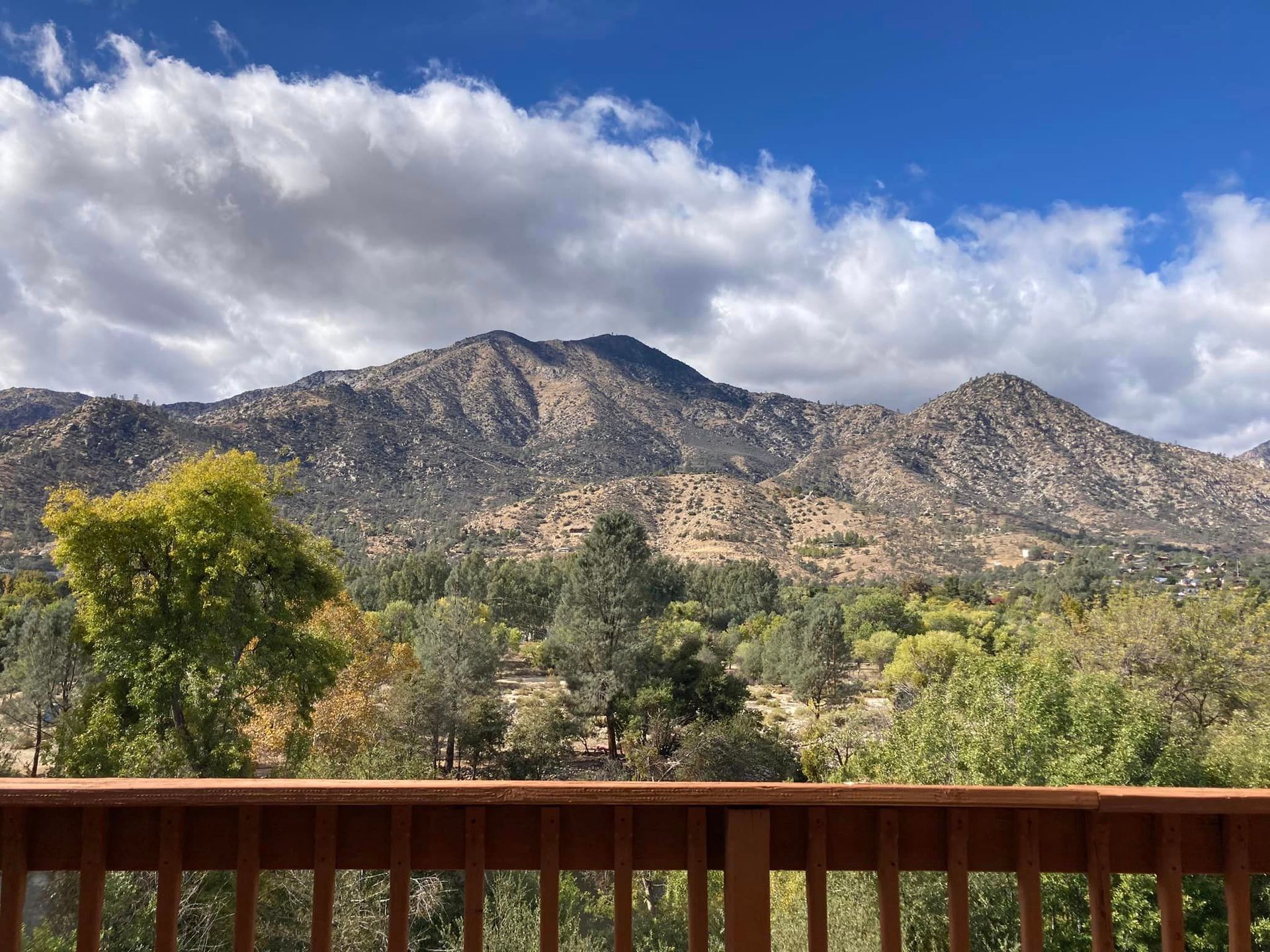 Mountain range viewed from a wooden deck, blue sky with white clouds, trees in foreground.