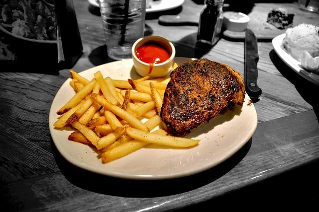 Steak with fries, tomato sauce, and a knife on a white plate on a wooden table.