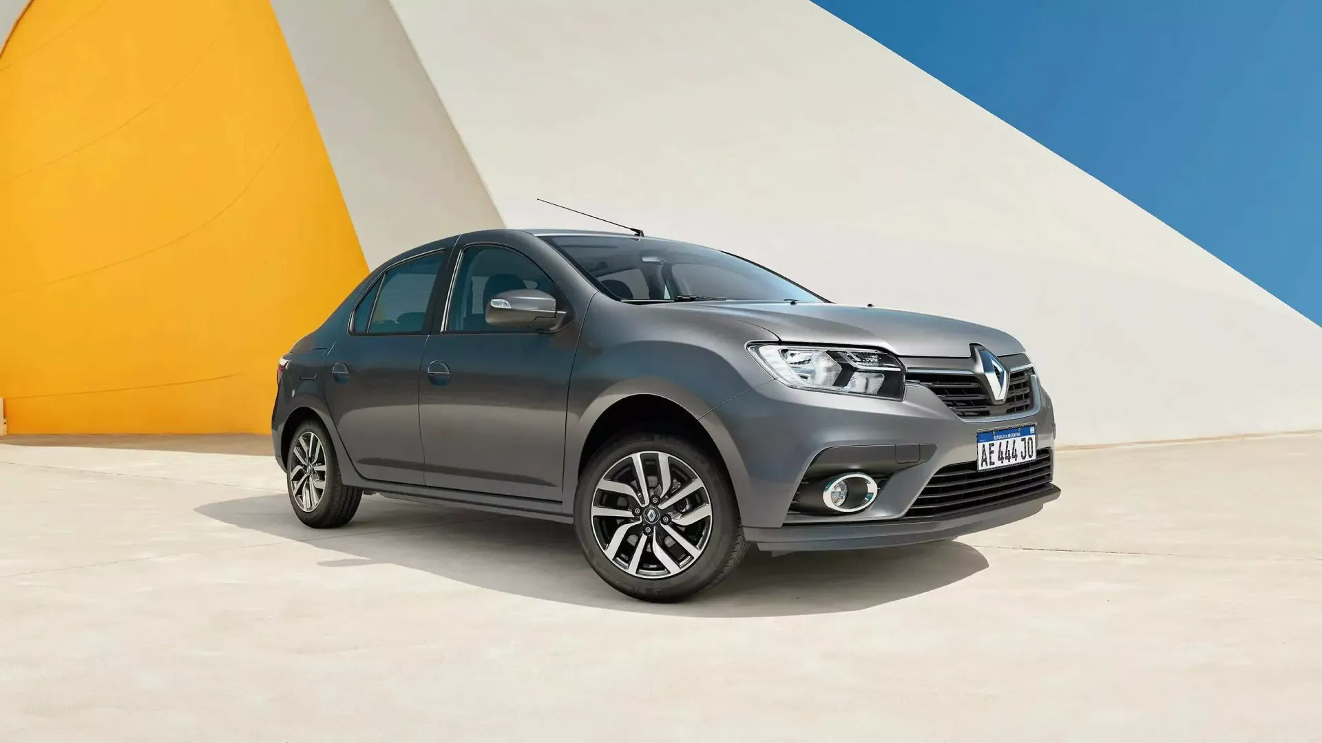 Un sedán Renault Logan de color gris estacionado frente a un fondo colorido.