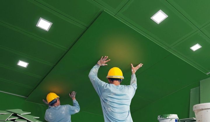 Workers in hard hats painting a bright green ceiling in a room with recessed lights