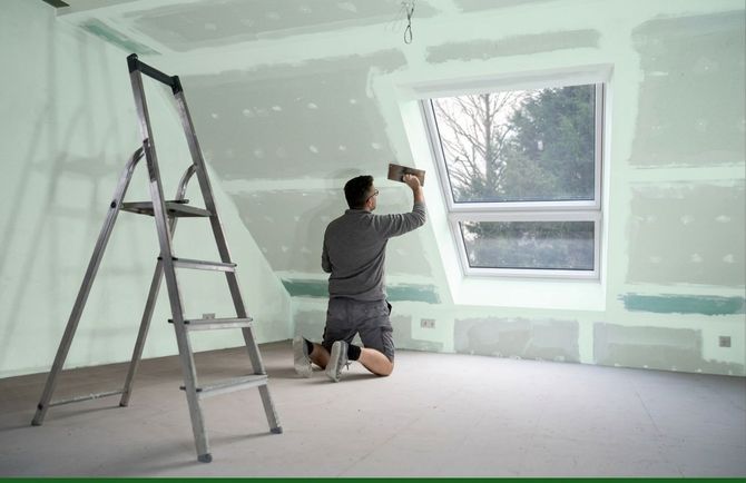 Person kneeling and painting a wall inside a room under renovation, with a ladder beside a large window.