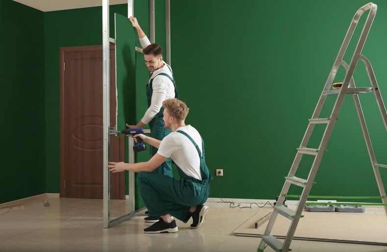 Two workers install drywall in a room with green walls and a ladder nearby.