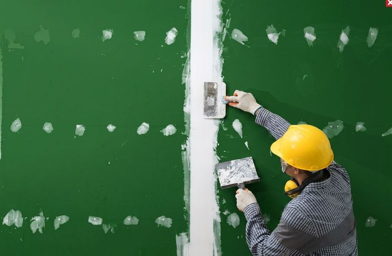 Construction worker in yellow hard hat smoothing white plaster on a green wall with a trowel