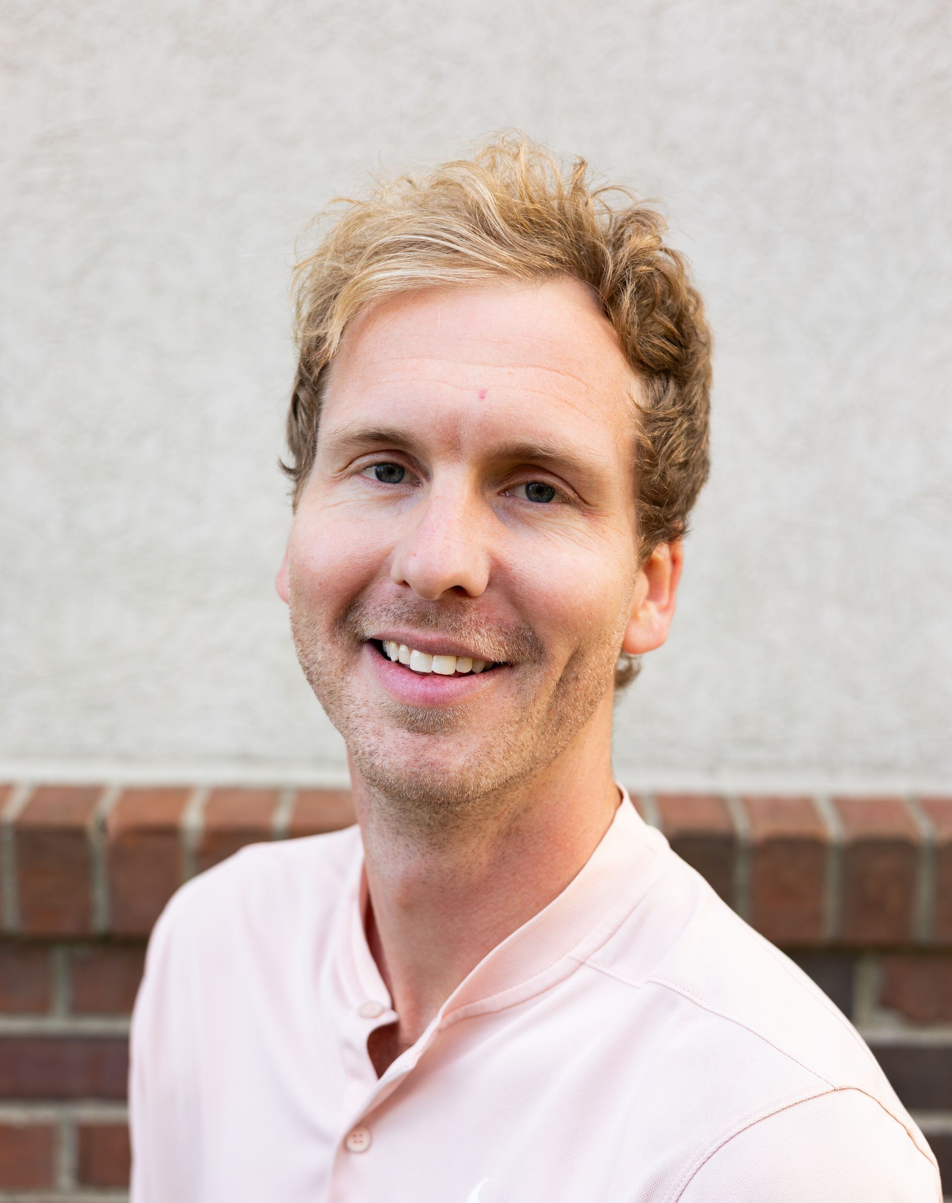 Man with light blonde hair and a pink shirt smiles in front of a brick wall.