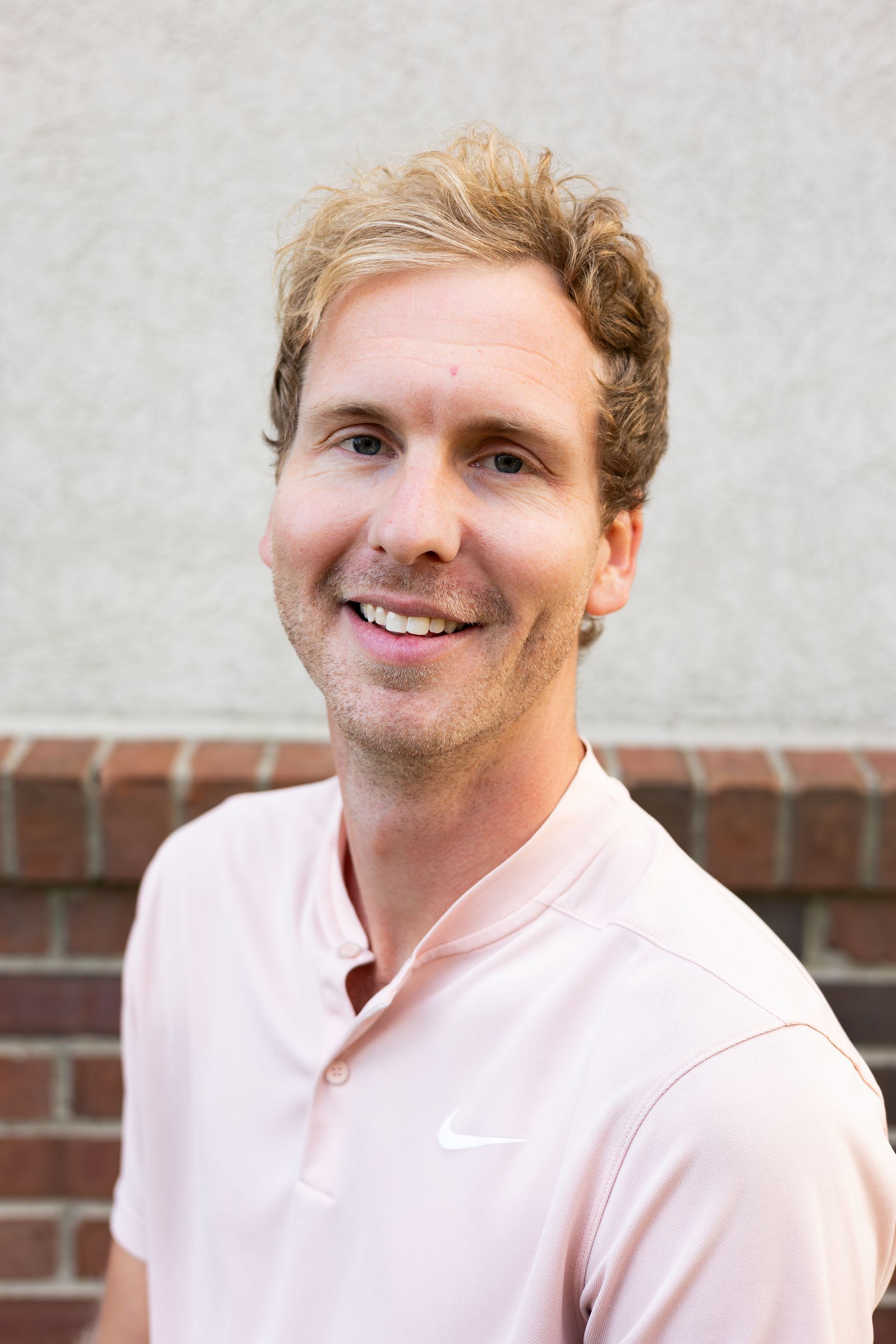 A man in a blue polo shirt is smiling in front of a brick wall.
