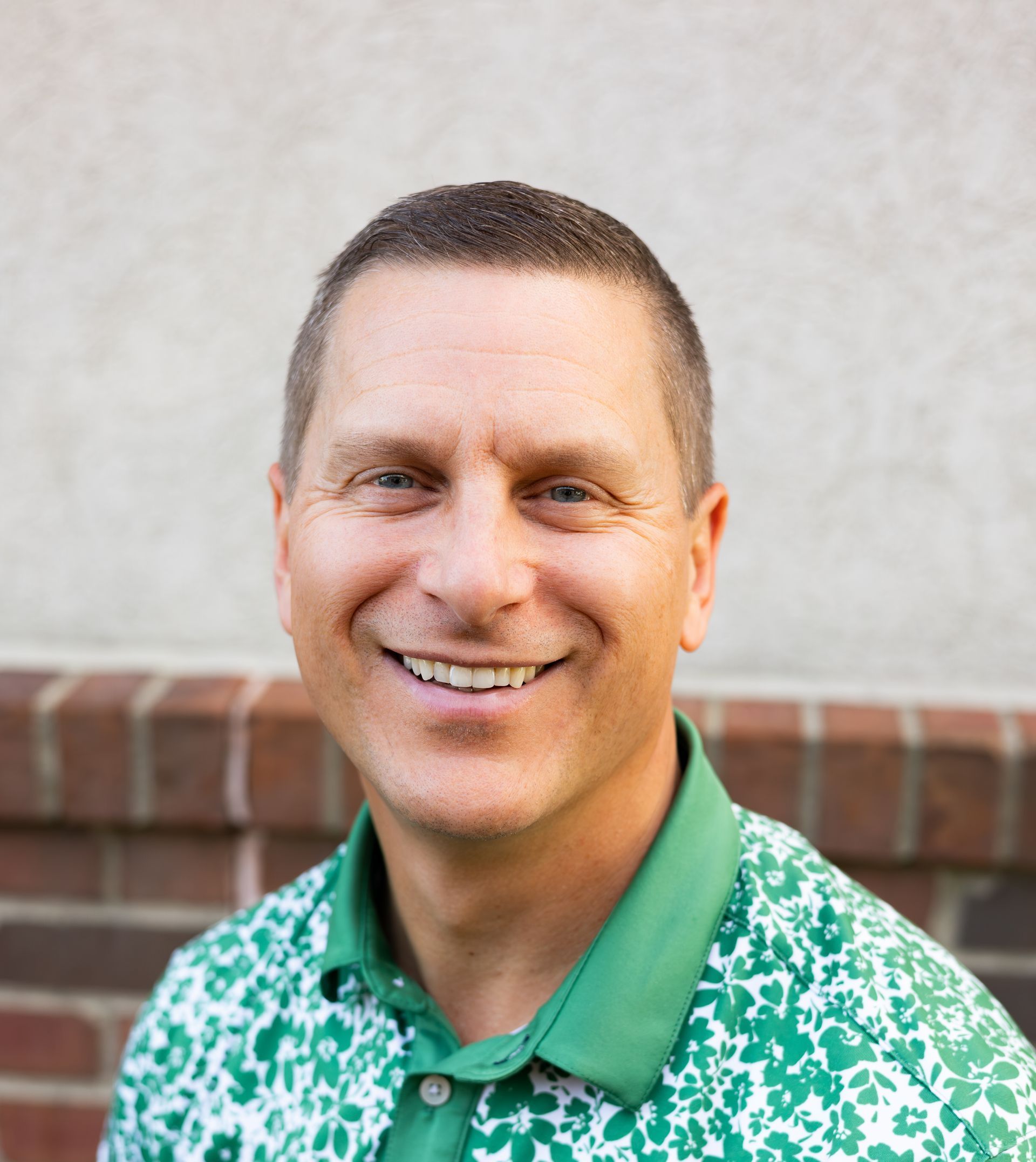 Smiling man with short brown hair, wearing a green floral polo shirt, against a brick wall.