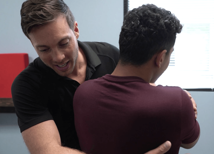 Chiropractor adjusting a man's back. Black shirted professional, smiling, with hands on a burgundy shirted patient. Office setting.