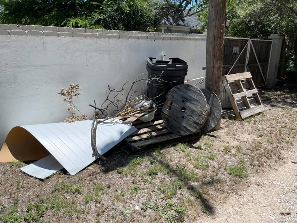 A pile of discarded items, including a light-colored sheet, a trash bin, wooden spools, and a pallet against a white wall.