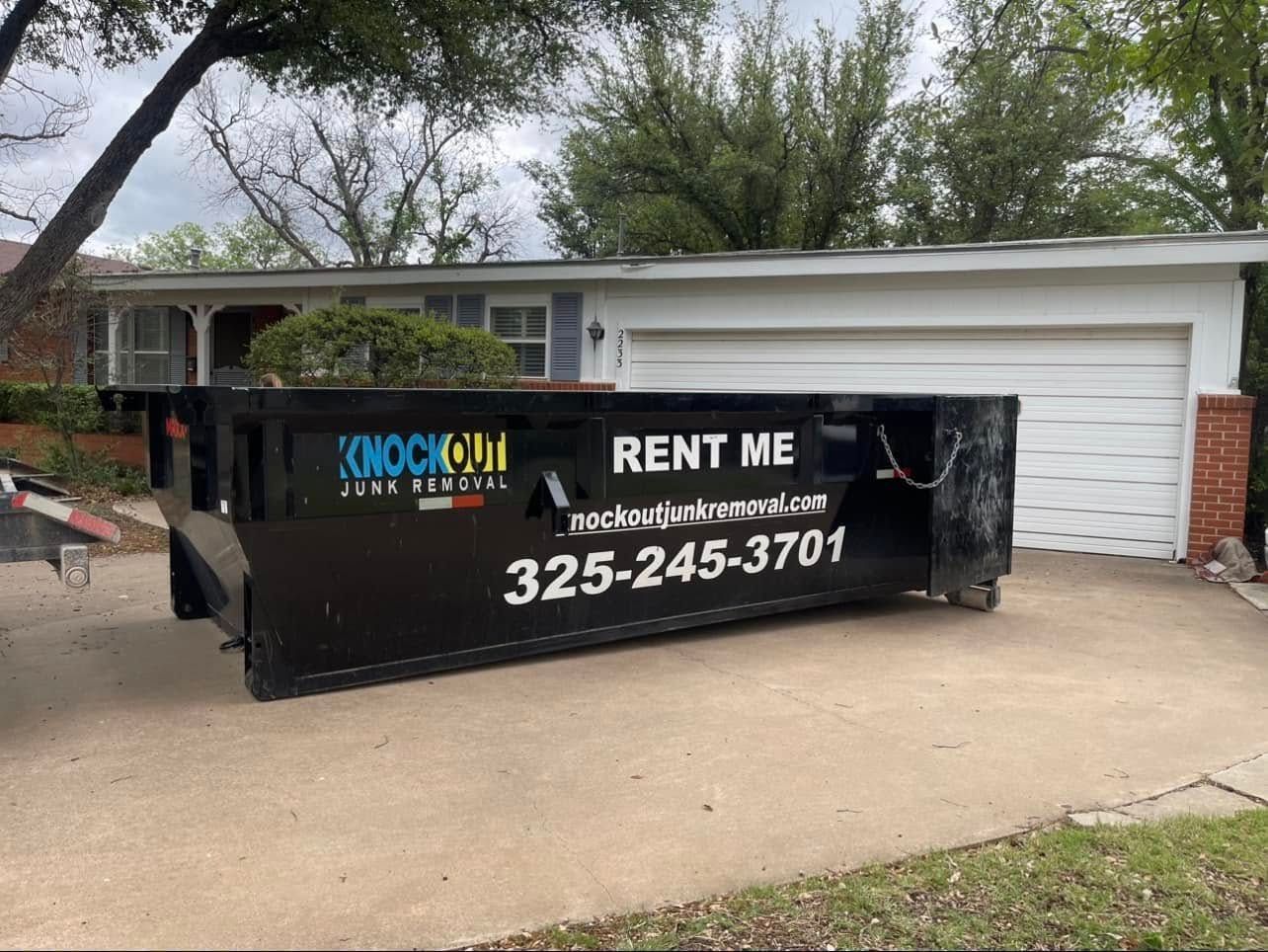 A black Knockout Dumpsters rental bin parked in a residential driveway in front of a house.
