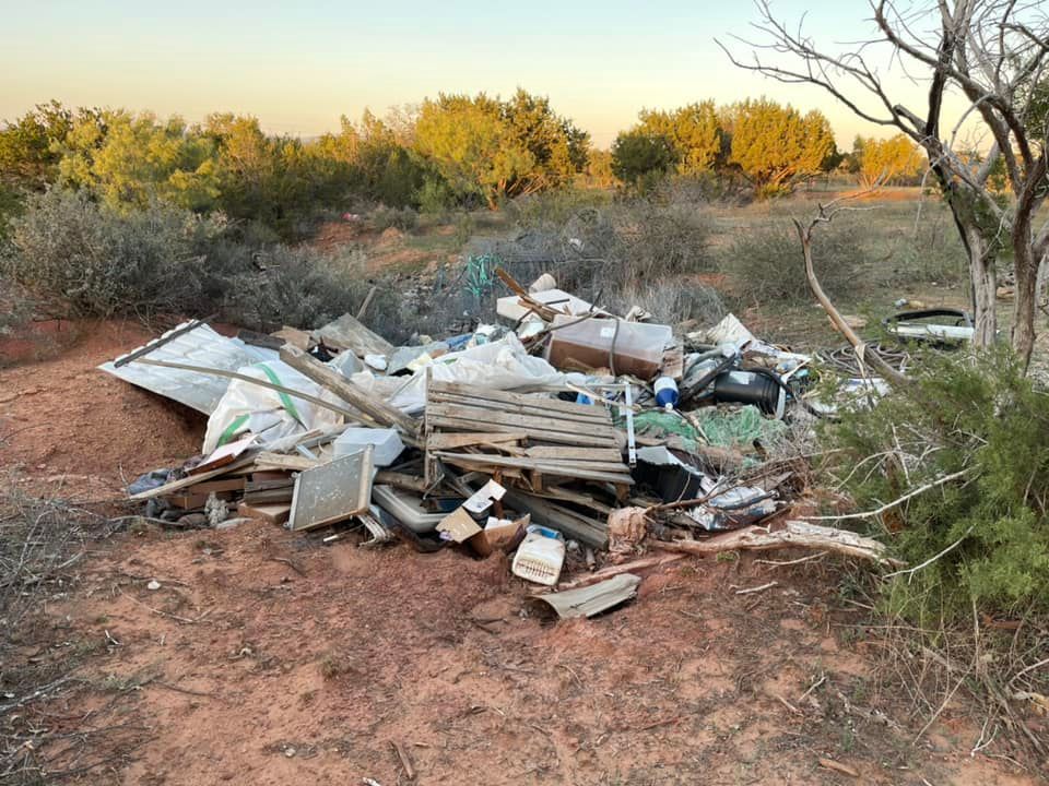 A large, messy pile of construction waste, scrap metal, and debris dumped in a dry, open field with sparse vegetation.
