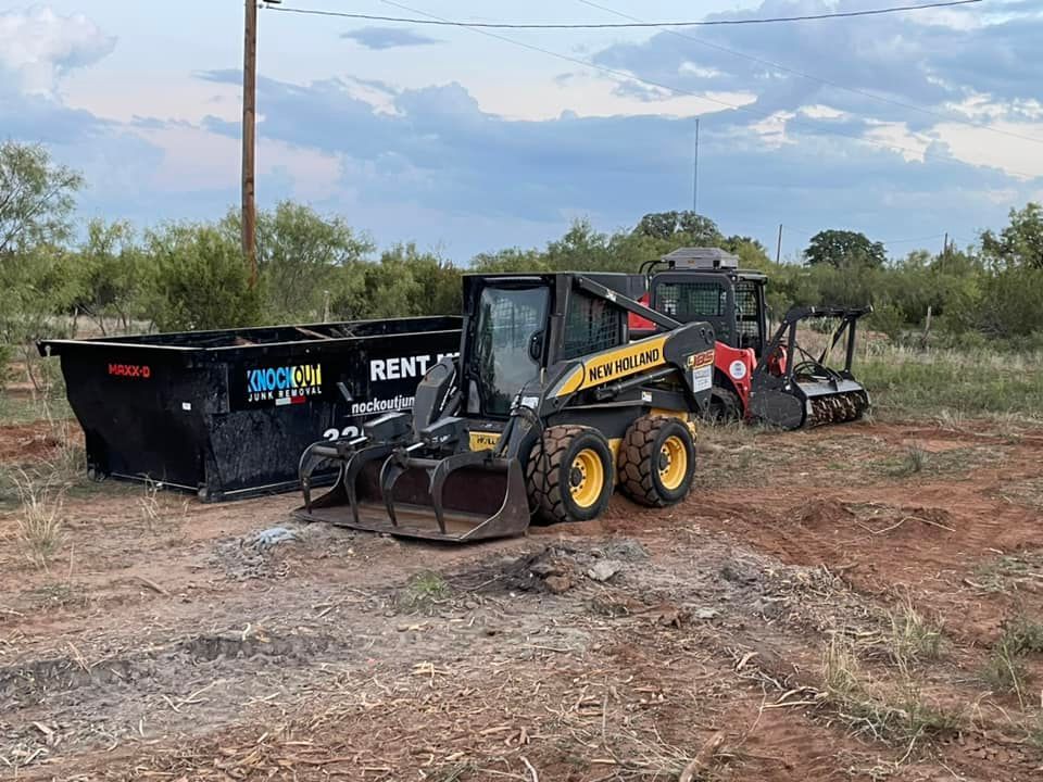 Two skid steer loaders and a black dumpster sit on a dirt lot with brush in the background under a cloudy sky.