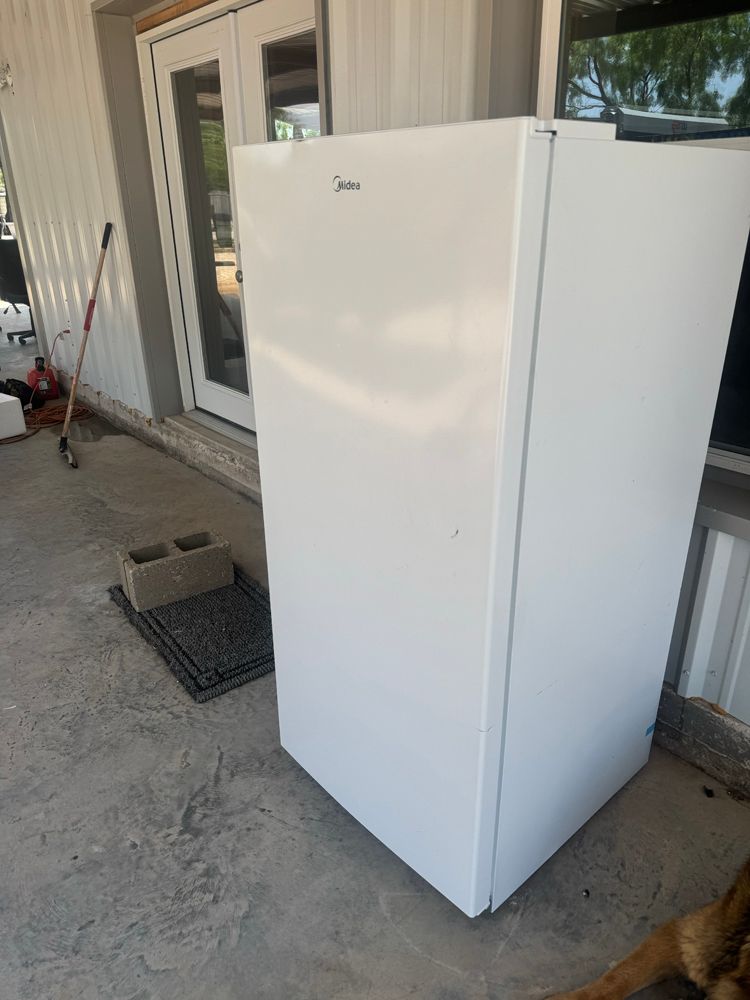 A white Midea upright freezer standing on a concrete patio outdoors near a doorway.