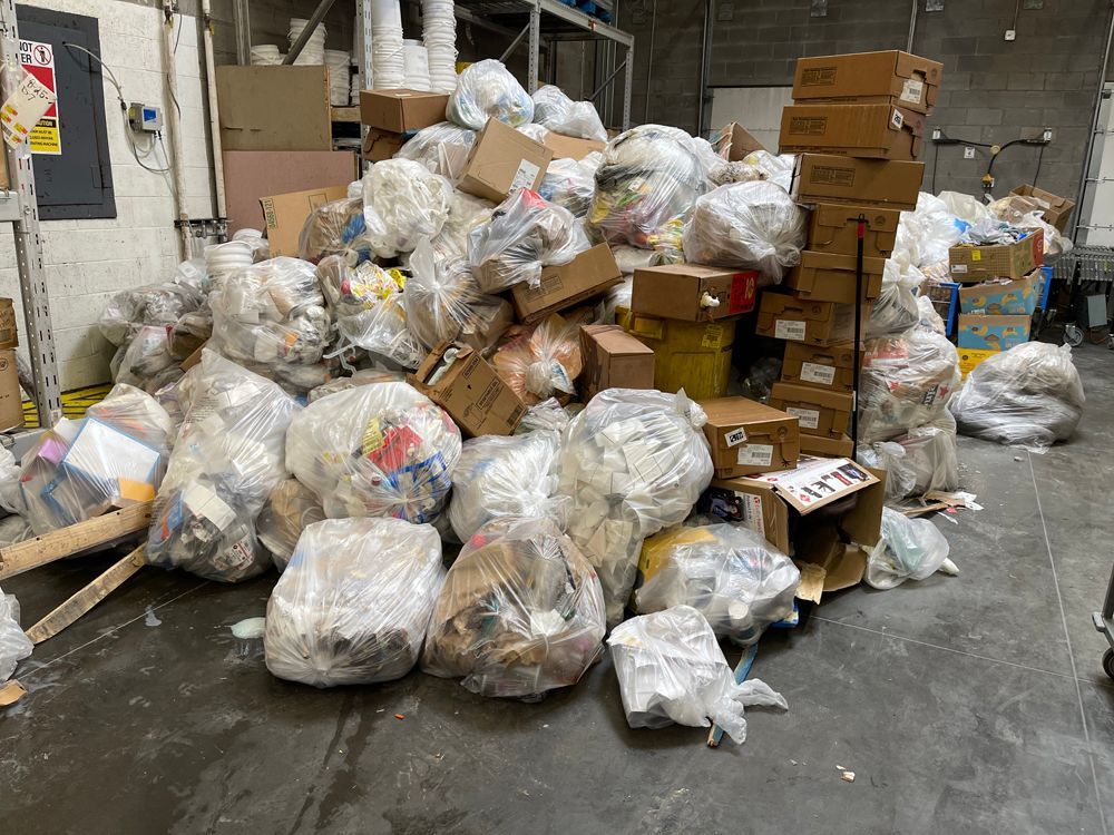 A large pile of clear plastic trash bags filled with waste and discarded cardboard boxes in a warehouse setting.
