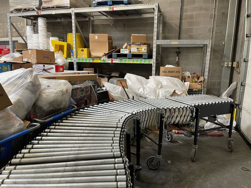 A curved metal conveyor belt sits in a storage area filled with shelves, cardboard boxes, and white plastic bags.