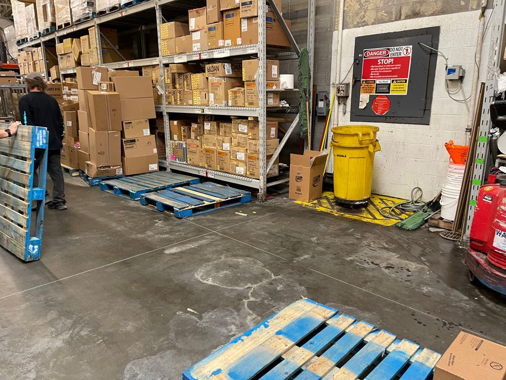A warehouse worker moves a blue pallet near storage shelves filled with cardboard boxes and a bright yellow trash bin.