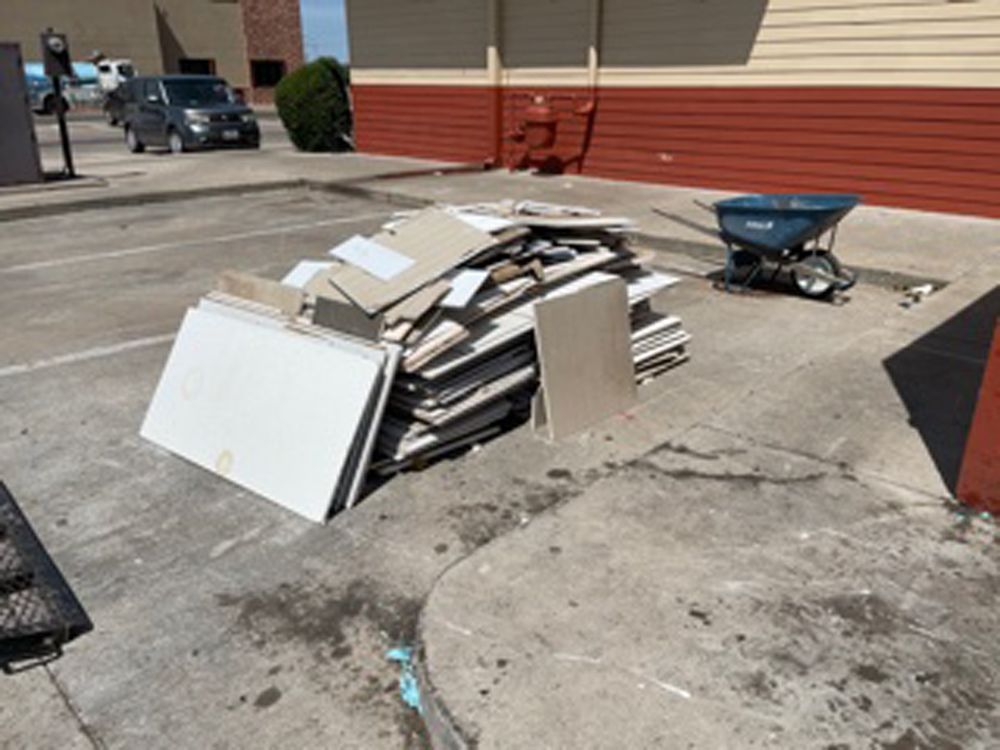 A pile of stacked construction waste and drywall boards sits on an outdoor concrete parking lot near a blue wheelbarrow.