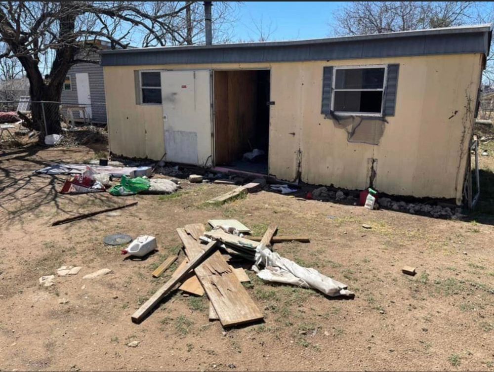 A dilapidated, light-colored mobile home with an open door stands in a yard littered with wood debris and trash.