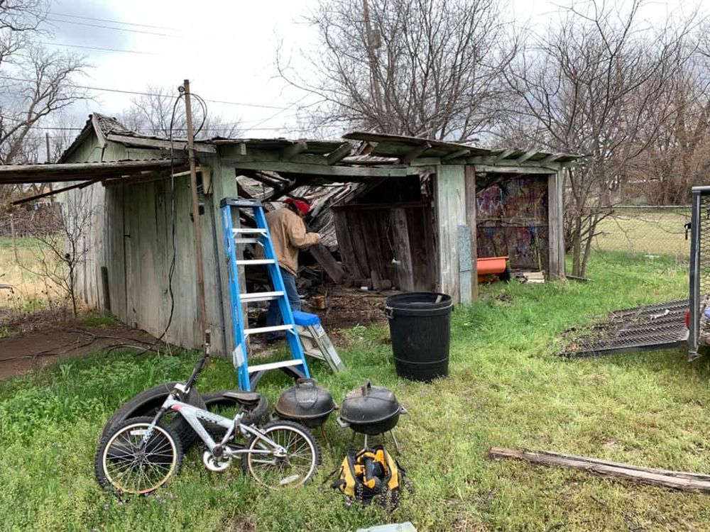 A person in a brown jacket works inside a dilapidated, partially collapsed wooden shed near a ladder, bike, and grills.