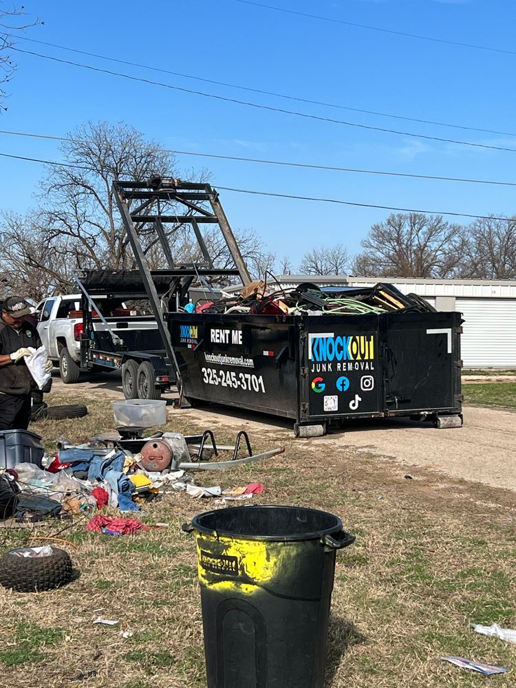 A dumpster labeled "Quick Out" hitched to a truck, with assorted debris on the ground in an outdoor, sunny setting.