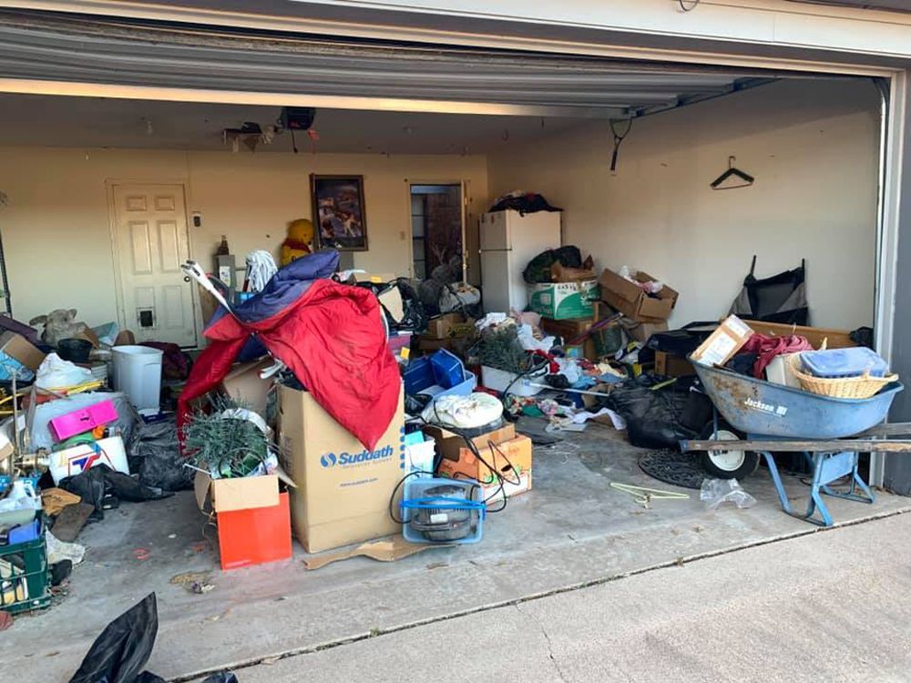 A cluttered, disorganized garage filled with cardboard boxes, trash bags, a blue wheelbarrow, and assorted household items.