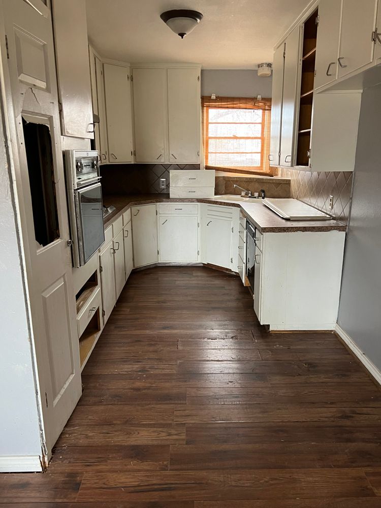 A galley-style kitchen with white cabinets, dark wood-look floors, a built-in oven, and a corner sink under a window.