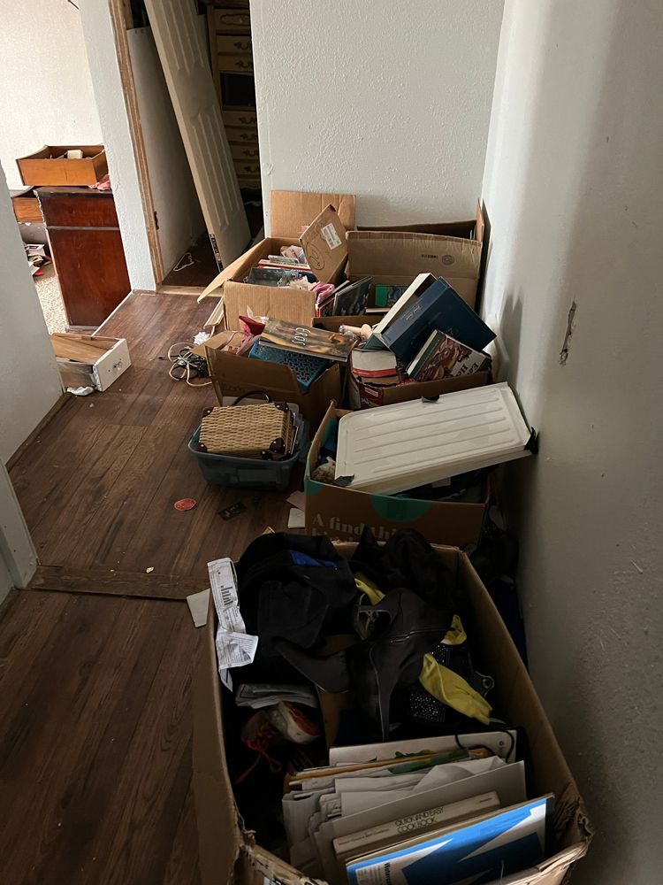 A cluttered hallway filled with cardboard boxes containing various household items, books, and miscellaneous objects.
